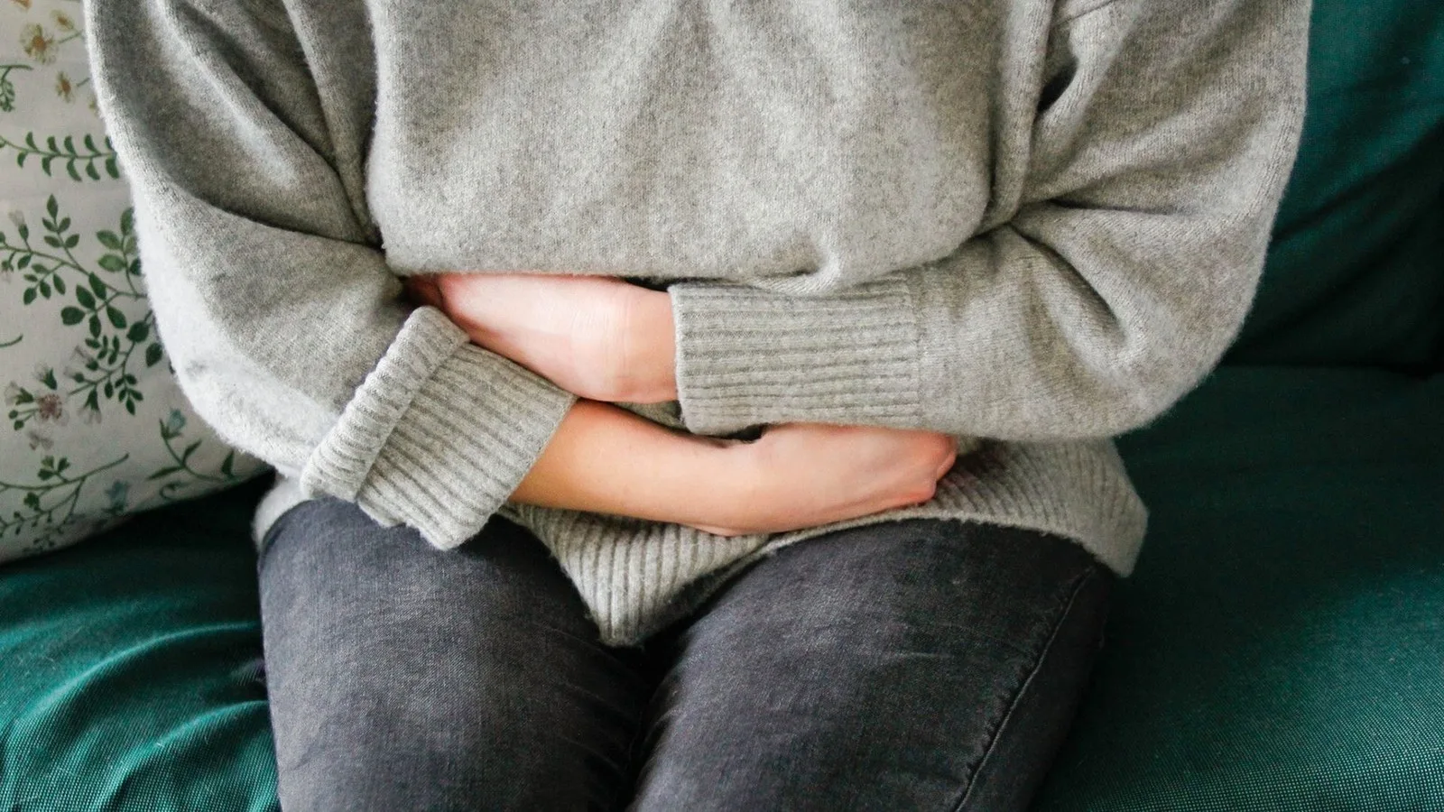 A woman wearing a gray sweater and blue jeans clutches her stomach.
