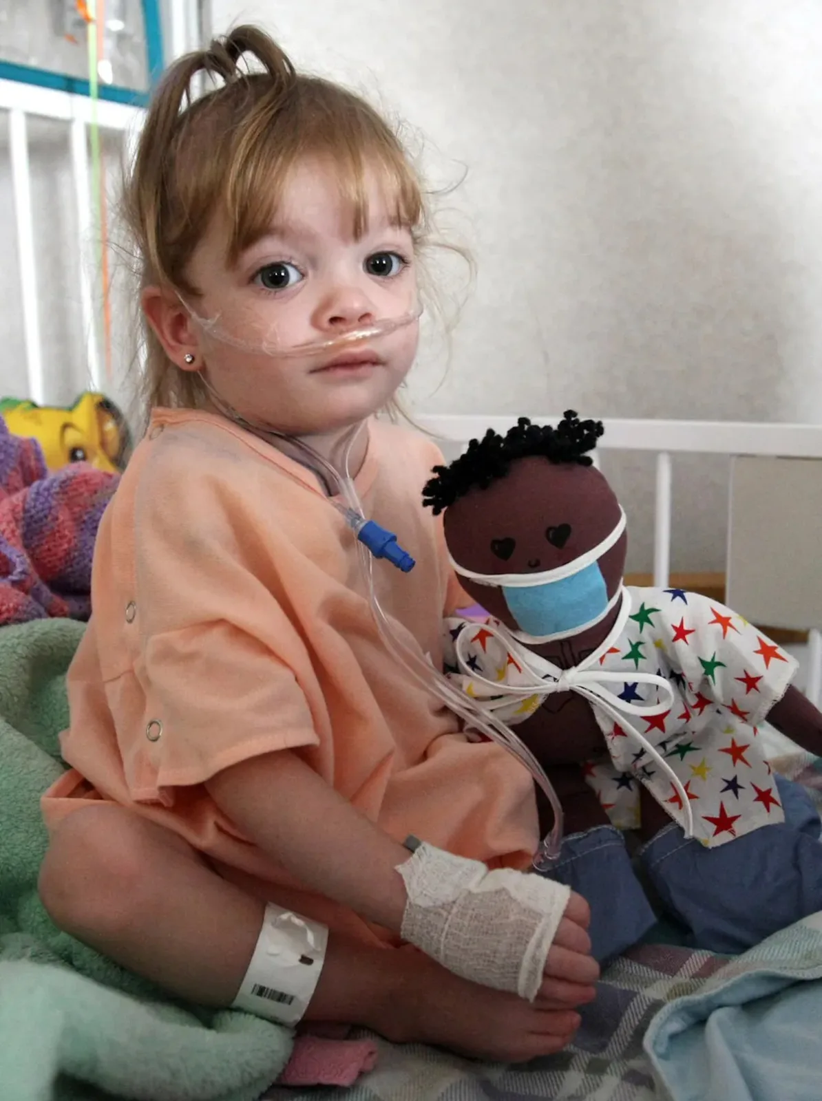 Child with nasal cannula sitting on bed, holding a doll wearing a surgical mask